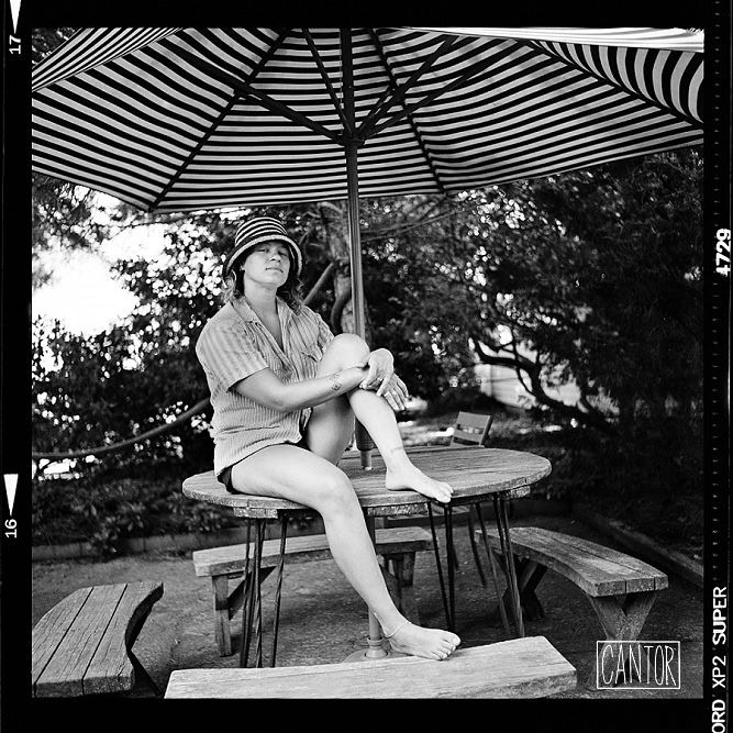 A woman sitting on a table under a striped patio umbrella outdoors, in a black and white photograph.