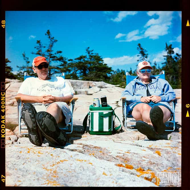 Two people relaxing on rocks by the coast with blue sky and trees in the background.